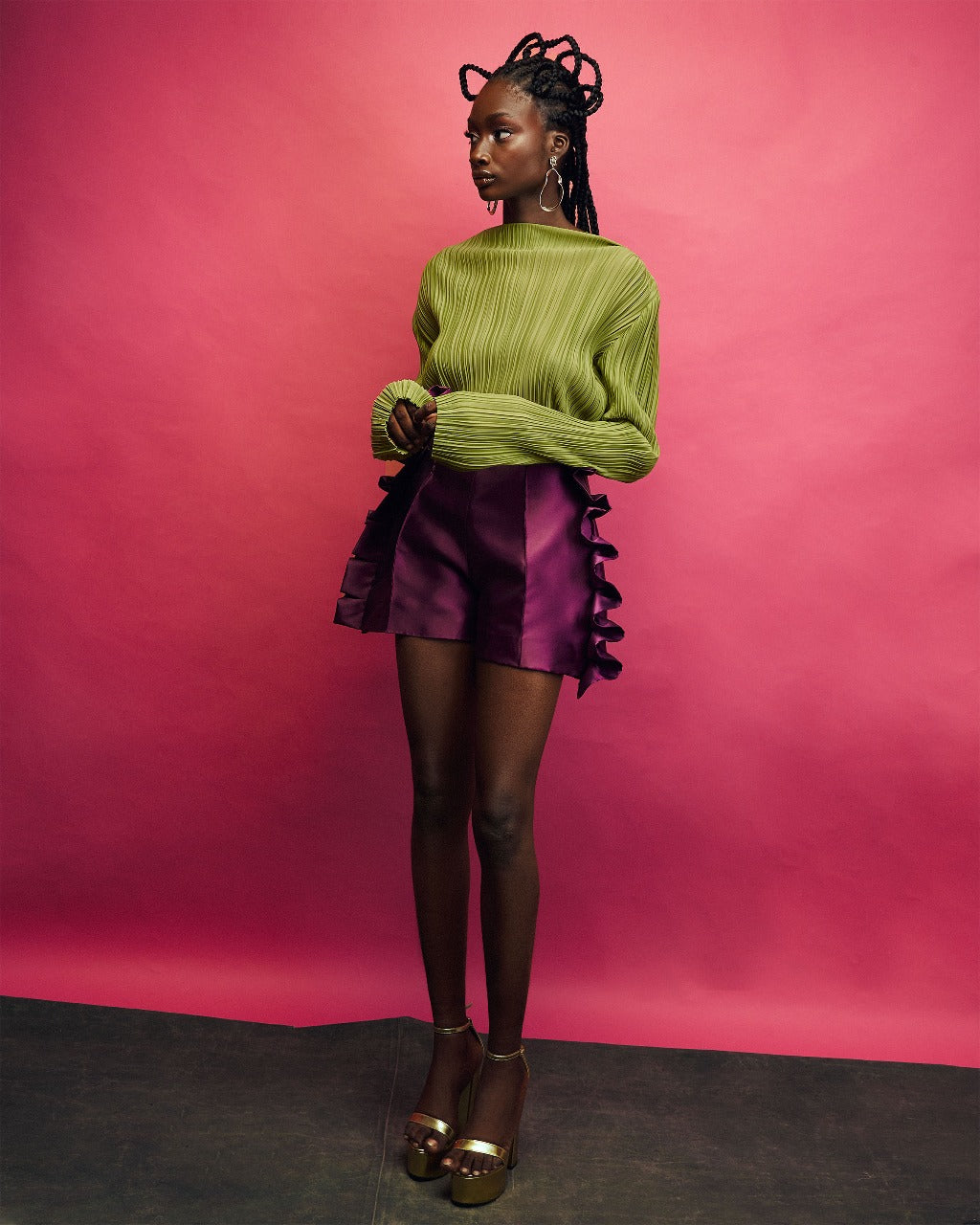 A model wearing a Chartreuse top and Aubergine colored shorts with ruffles in a red room
