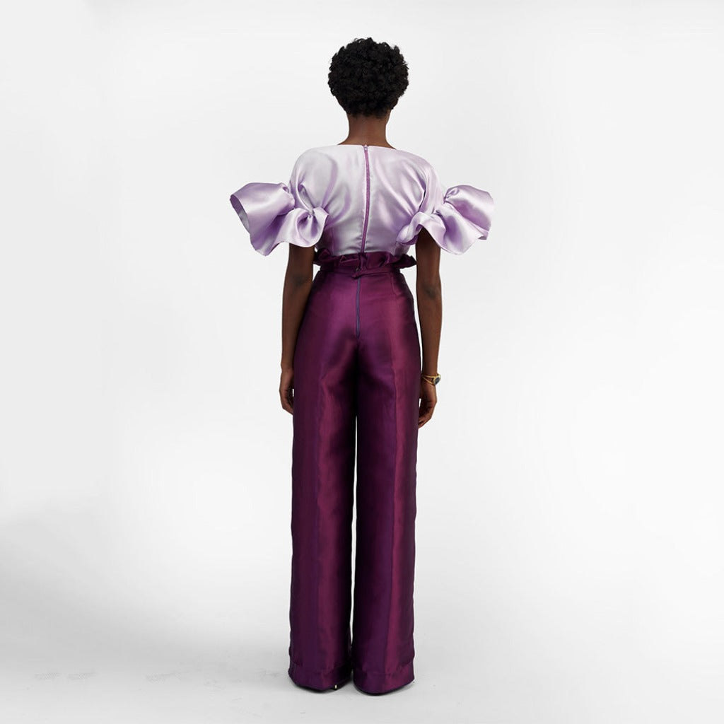 The back of a model wearing aubergine colored pants with a purple top in a white room