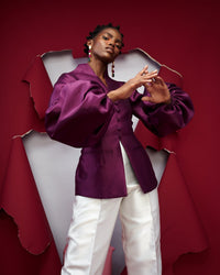 A close-up of a model posing wearing aubergine colored silk satin jacket with white pants in a red setting
