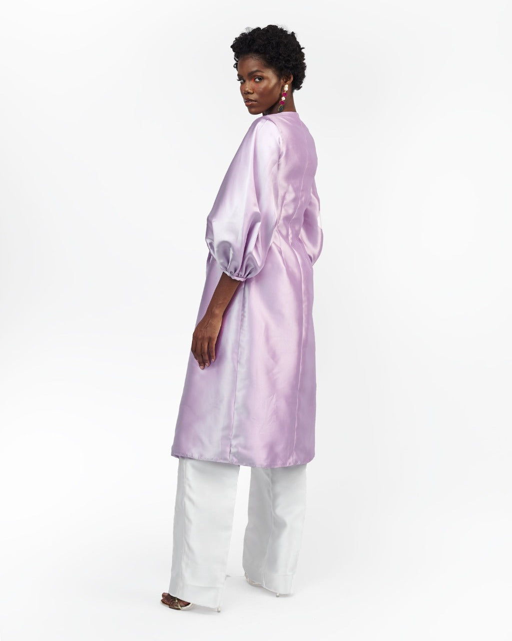 The back of a model in a Lilac silk satin jacket with puff sleeve and a White straight-cut pant in a white room