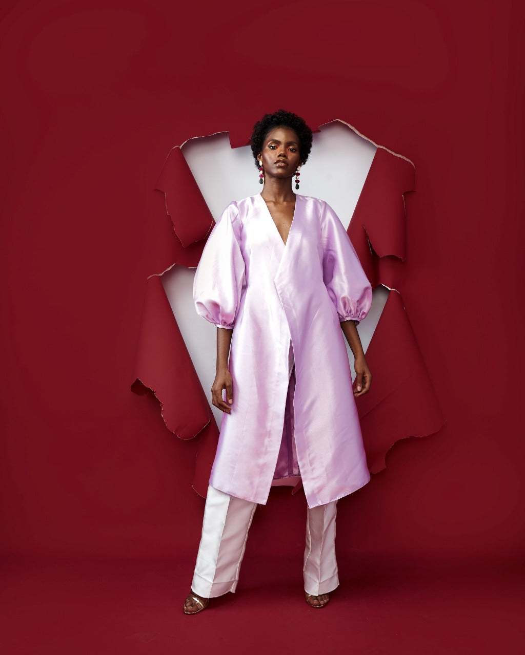 A model in a Lilac silk satin jacket with puff sleeve and a White straight-cut pant in a red room