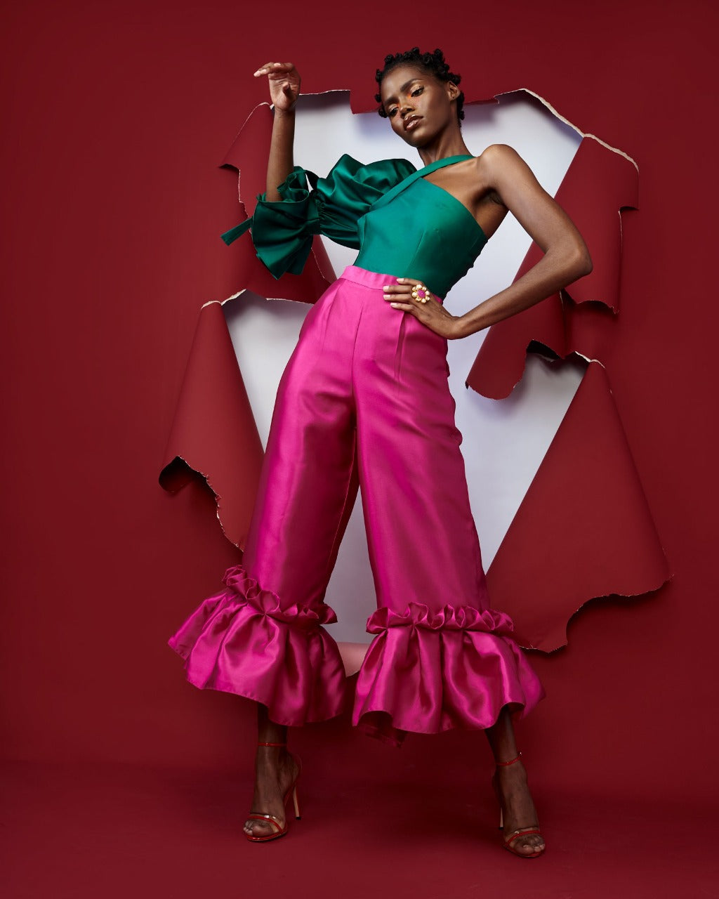 A model posing wearing a silk satin green top and magenta pants in a red room