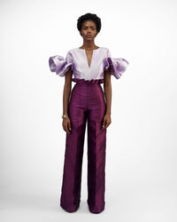 A model wearing aubergine colored pants with a purple top in a white room