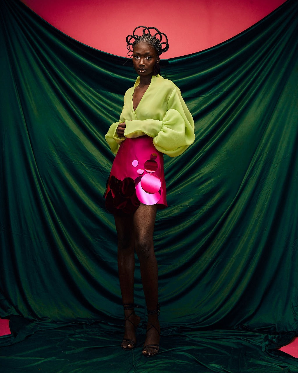 A model wearing a Chartreuse top and a Magenta high waist mini skirt with sequins embellishment in front of a green sheet