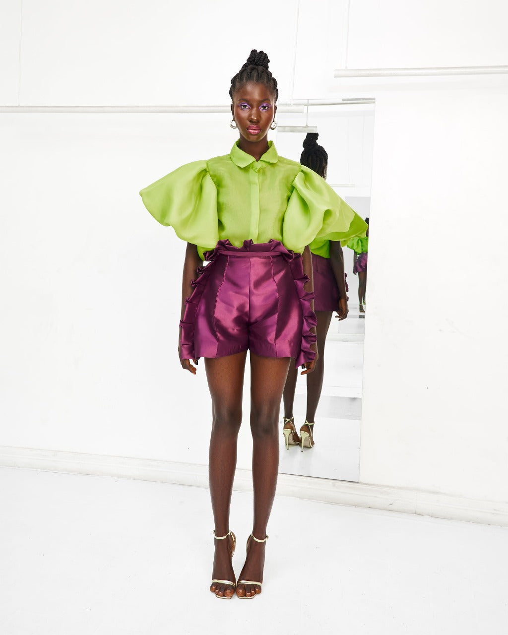 A model wearing a Chartreuse top and Aubergine colored shorts with ruffles in a white room