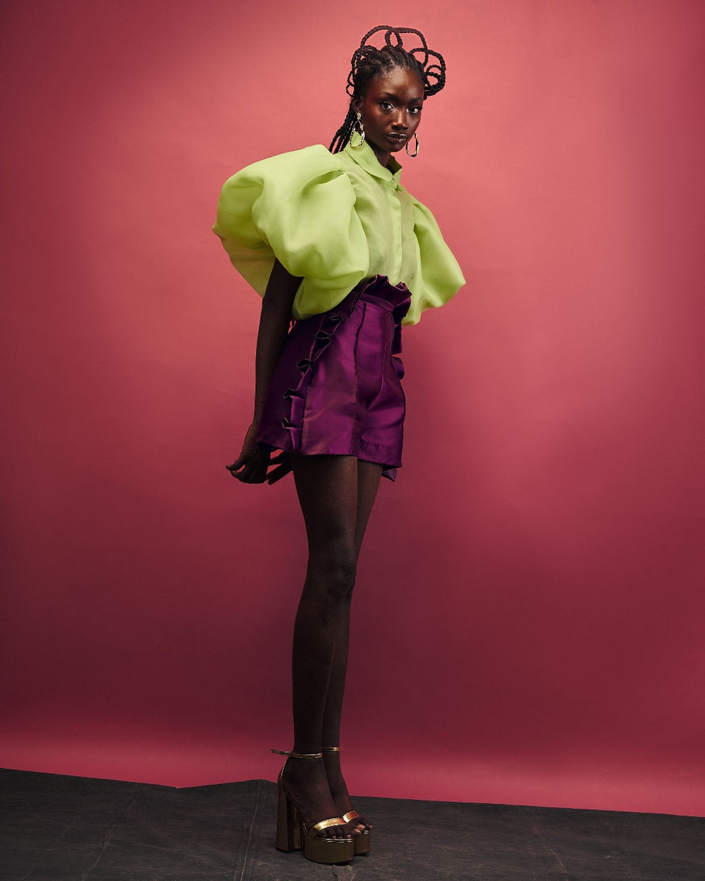 A model wearing a Chartreuse top and Aubergine colored shorts with ruffles in a red room