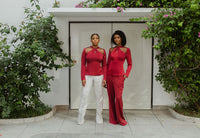 Two models wearing a burgundy top and white and burgundy pants in front of a white house