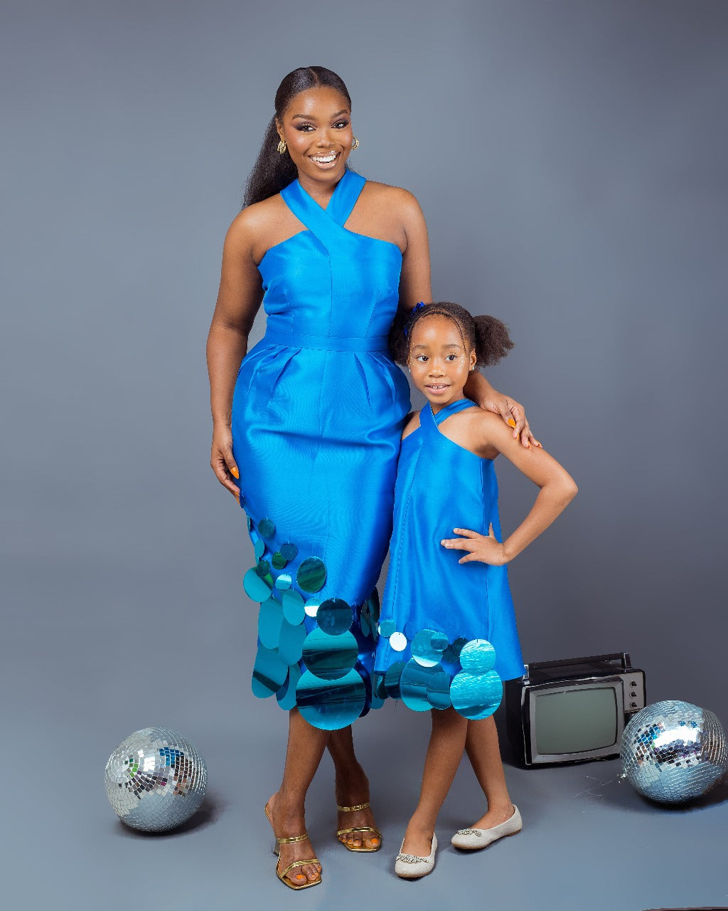 A girl wearing a Turquoise A-line dress with a crisscross neckline and sequins embellishment, and a woman wearing a Turquoise top and skirt with crisscross neckline and pleat details at the waist with sequins embellishment