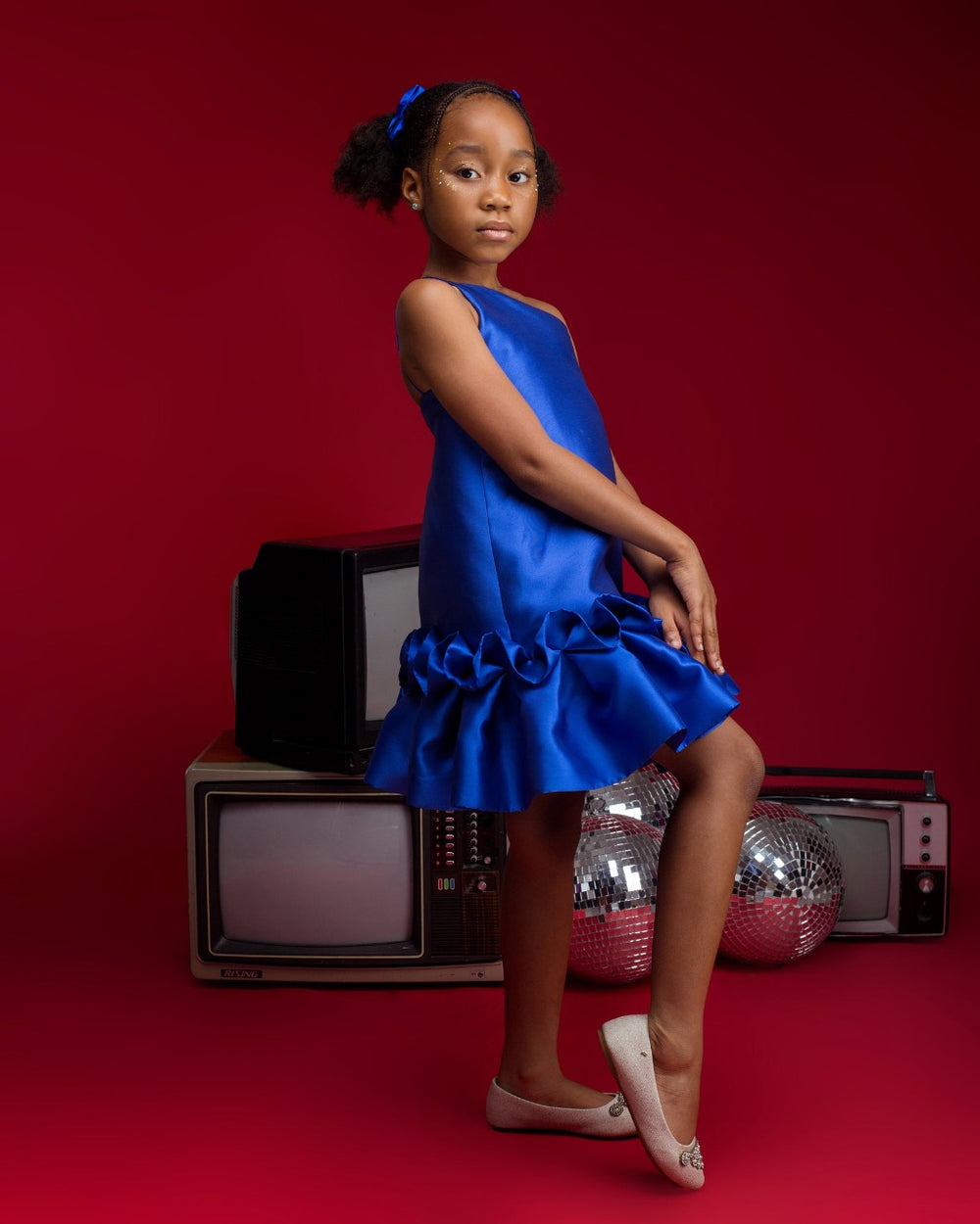 A kid model wearing a one-shoulder blue dress with drop waist ruffle detailing in a red room