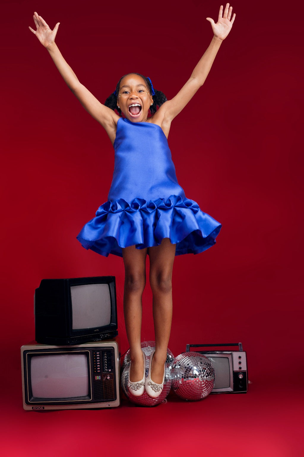 A smiling kid model wearing a one-shoulder blue dress with drop waist ruffle detailing in a red room