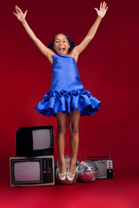 A smiling kid model wearing a one-shoulder blue dress with drop waist ruffle detailing in a red room