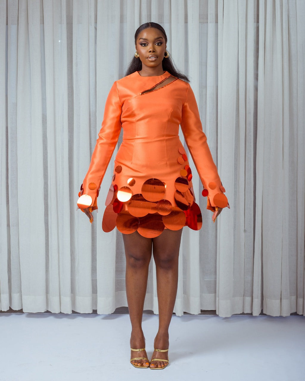 A model wearing an Orange mini dress with side cut-out detail at the neckline and embellished with sequins at the hem and sleeves in front of white curtains