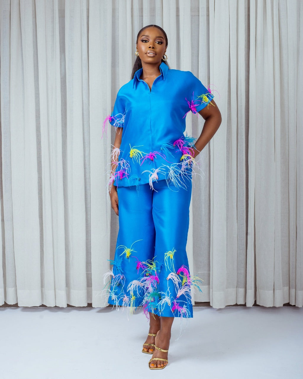 A model wearing a Turquoise shirt with a short sleeve attached with ostrich feathers and a Turquoise pants with ostrich feathers embellishments in front of white curtains
