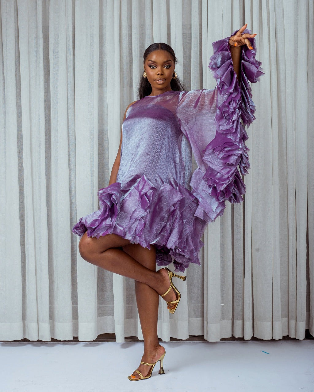 The side of a model wearing a Purple asymetric sleeve mini dress with ruffles and silk slip lining in front of white curtains