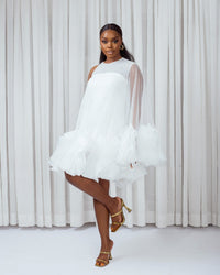 A model wearing a White asymetric sleeve mini dress with ruffles and silk slip lining in front of white curtains