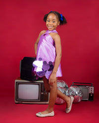 The side of a kid model wearing a lilac dress with a crisscross neckline and sequins embellishment at the hem in a red room