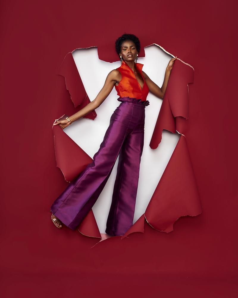 A model posing in a red room wearing purple silk satin pants with an orange top 